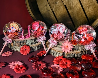 May include: Four clear glass ball ornaments with pink and red accents. Each ornament features a different anime character. The ornaments are on a wooden surface with red flowers and red glass stones.