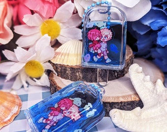 May include: Two cartoon characters are encased in clear blue resin with a silver chain. The resin is filled with small blue fish. The characters are a girl with pink hair and a boy with blonde hair. The resin is shaped like a popsicle.