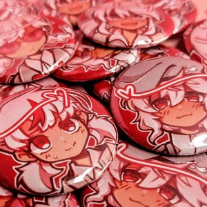 May include: A collection of red and white button pins featuring anime-style characters with pink hair and red eyes. The characters are wearing white shirts and have different expressions.