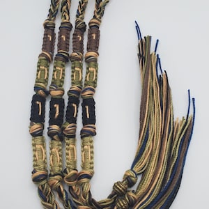 Pode incluir: Três cordões trançados com borlas, cada um com uma série de contas com letras hebraicas. Os cordões são em tons de marrom, verde e azul.