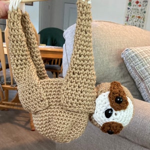 May include: A crocheted sloth hanging from a wooden dowel. The sloth is brown with white accents and has a brown face with black eyes.
