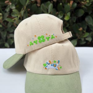 May include: Two beige corduroy baseball caps with olive green brims. The top cap features embroidered green four-leaf clovers and small colourful dots. The bottom cap displays an embroidered illustration of a white cat with blue spots and flowers.