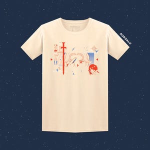 May include: Cream-colored t-shirt with a graphic design featuring a red sword, anime-style eyes, and a red-haired character. The text "POPOALU" is printed on the sleeve and near the graphic. The shirt has a crew neck and short sleeves.
