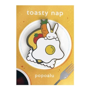Può includere: Uno spillo in smalto bianco con un coniglio dei cartoni animati che indossa un uovo fritto come coperta. Il coniglio tiene una carota e un pomodoro. Lo spillo è su uno sfondo giallo con il testo "toasty nap" e "popoalu".