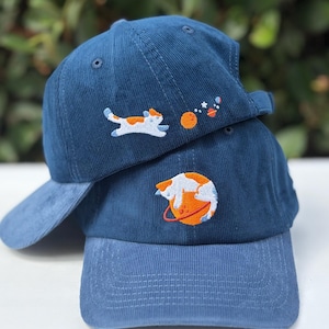 May include: Two navy blue corduroy baseball caps featuring embroidered space-themed designs. The top cap displays a cat floating near a planet and stars. The bottom cap features a cat curled around a large orange planet with a red ring.