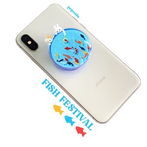 May include: A blue and white phone pop socket with a cartoon fish pond design. The pop socket features a white rabbit and a yellow cat fishing in the pond. The text "FISH FESTIVAL" is printed on the phone case below the pop socket.