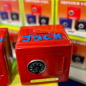 May include: Red metal toy safe with a combination lock and the word "Jack" written in blue letters on the front. The safe is a piggy bank and is part of a collection of toy safes.