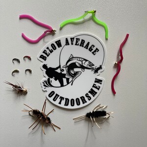 May include: A white sticker with the text "Below Average Outdoorsmen" and a silhouette of a person fishing. The sticker is surrounded by various fishing lures, including green, pink, and red worm-shaped lures, and black and brown flies.
