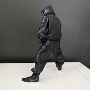 May include: A black hooded jumpsuit with matching black pants and black high-top sneakers with a red swoosh logo. The sneakers have a white sole. The figure is posed with a slight bend in the knees.