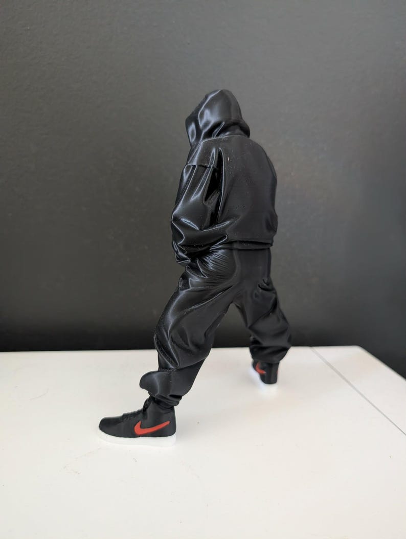May include: A black hooded jumpsuit with matching black pants and black high-top sneakers with a red swoosh logo. The sneakers have a white sole. The figure is posed with a slight bend in the knees.