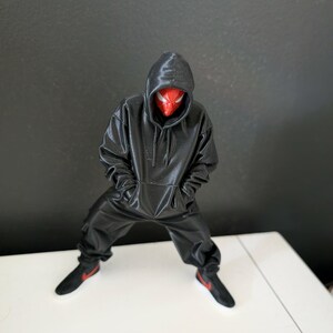 May include: A black figure in a hooded sweatshirt and matching pants, with a red and black mask. The figure is wearing black sneakers with red accents. The figure is standing on a white surface against a dark gray background.