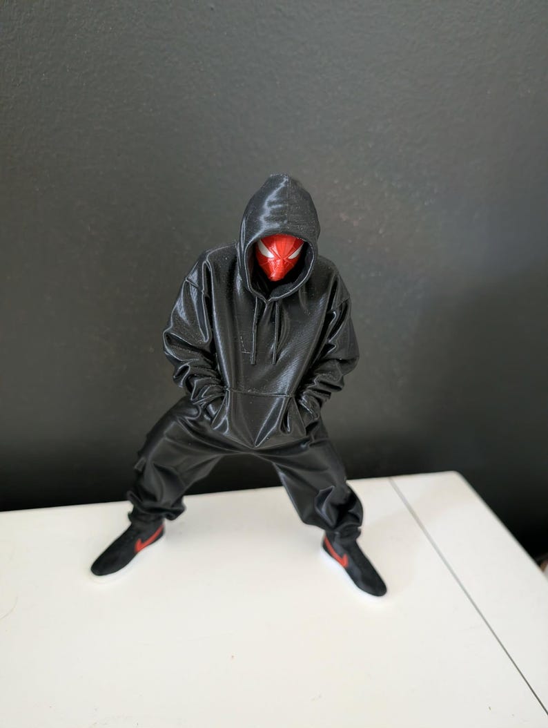 May include: A black figure in a hooded sweatshirt and matching pants, with a red and black mask. The figure is wearing black sneakers with red accents. The figure is standing on a white surface against a dark gray background.