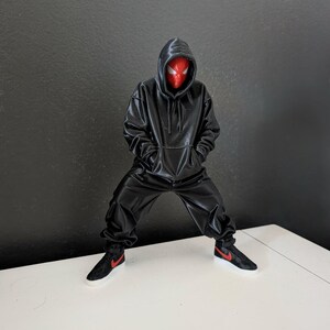 May include: A figurine of a superhero wearing a black hoodie and matching pants, with a red mask. The figure is posed with its legs apart and hands in the pockets of the hoodie. Black sneakers with red accents complete the outfit.