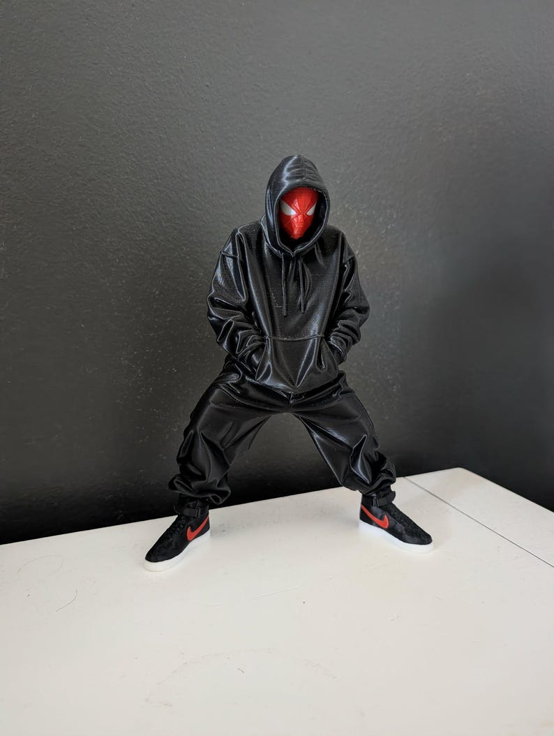 May include: A figurine of a superhero wearing a black hoodie and matching pants, with a red mask. The figure is posed with its legs apart and hands in the pockets of the hoodie. Black sneakers with red accents complete the outfit.