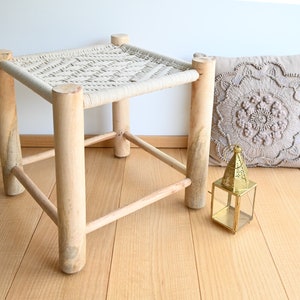 Op de afbeelding: Een natuurlijke houten kruk met een geweven bovenkant en een decoratief kussen en een lantaarn op de vloer.