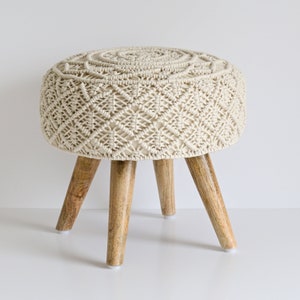 Op de afbeelding: Een witte macramé kruk met een ronde bovenkant en vier houten poten. De kruk heeft een geweven ontwerp en is perfect om elke kamer een bohemian touch te geven.
