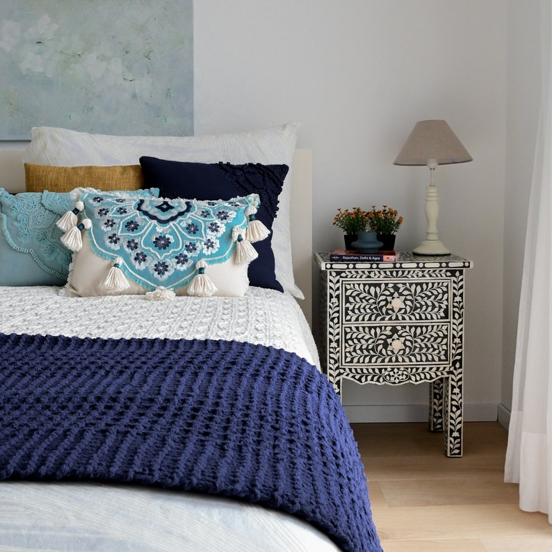 May include: A white bed with a blue and white patterned throw blanket and a blue and white patterned pillow. A black and white nightstand with two drawers is next to the bed. A lamp with a white shade is on the nightstand. The nightstand has a book on it that says "Aquamarine, Delhi & Agra".