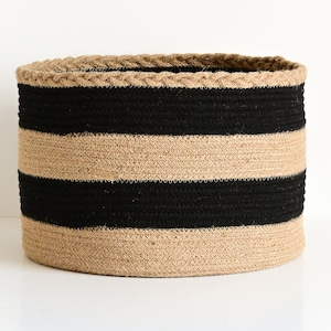 Op de afbeelding: Een grote, ronde, gestreepte opbergmand gemaakt van natuurlijke jute touw met zwarte strepen. De mand is geweven en heeft een gevlochten touwrand.