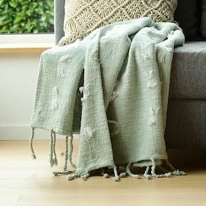 Peut inclure: Une couverture en jet douce de couleur vert clair avec un tissage texturé et des franges blanches. La couverture est drapé sur un canapé avec un coussin de jet de couleur crème.