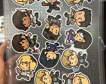 May include: A collection of fourteen die-cut stickers featuring chibi-style cartoon characters with various expressions, poses, and accessories, arranged on a transparent adhesive sheet with a printed grid pattern background.