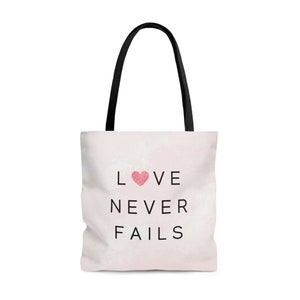 May include: A white tote bag with black handles and a pink watercolour background. The text "LOVE NEVER FAILS" is printed in black with a pink heart in the middle of the word "LOVE".