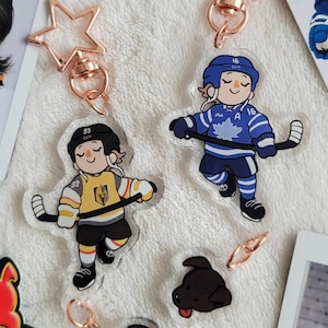 Puede incluir: Colección de llaveros y cromos con temática de hockey. Los llaveros presentan jugadores de hockey de dibujos animados con uniformes de equipo, con cierres en forma de estrella. Incluye una foto y una pegatina con un jugador de hockey. La combinación de colores incluye azul, amarillo y negro.