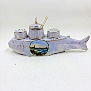 May include: A pearlescent ceramic fish-shaped condiment set featuring a central lidded mustard pot with a small spoon, flanked by two perforated shakers. The fish body displays a circular landscape decal with the text Nordseebad Cuxhaven.