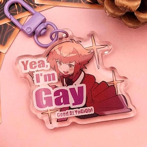 Peut inclure: Un porte-clés en acrylique transparent avec un fermoir violet. Le porte-clés présente un personnage de style anime avec le texte "Yea, I'm Gay" et "Good At YuGiOh!" en rose et blanc. Le personnage a les cheveux roux et porte une tenue rouge.