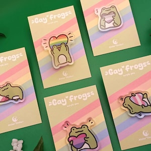 May include: Five colorful cards with rainbow stripes and the text "Gay Frogs" printed on them. Each card features a different frog design with a heart and a rainbow. The cards are for sale as acrylic pins.