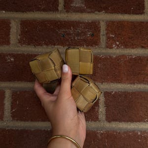 May include: Three small, cube-shaped objects crafted from woven light brown strips of natural material. Each cube features a tight, interlocking grid pattern with visible edges and corners, displaying a consistent tan color and organic texture throughout.