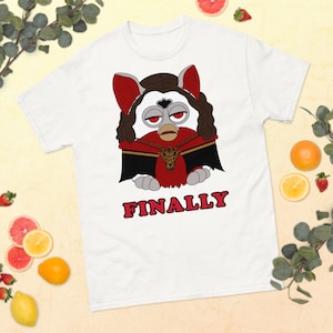 May include: White t-shirt with a cartoon image of a vampire Furby wearing a red cape and a gold necklace. The text "FINALLY" is printed in red below the image.