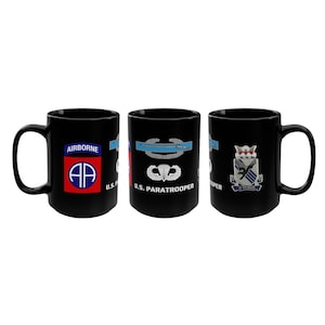 May include: Three black ceramic mugs with military-themed designs. One mug features a blue and red emblem with the word "AIRBORNE" and "AP". Another mug displays a parachute and the text "U.S. PARATROOPER". The third mug has a crest.