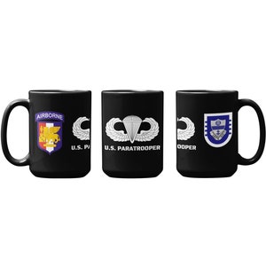 May include: Three black ceramic mugs with white designs. The mug on the left has a purple and gold crest with the words "Airborne" and "U.S. Paratrooper". The middle mug has a white parachute with wings and the words "U.S. Paratrooper". The mug on the right has a blue and white crest with the word "Paratrooper".