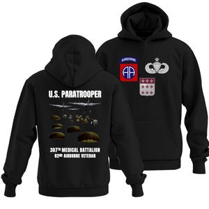May include: Black hooded sweatshirt featuring the text "U.S. Paratrooper" with an image of paratroopers and planes. The front displays military patches, including an Airborne patch and a parachute badge. The back of the sweatshirt also has the text "307th Medical Battalion 82nd Airborne Veteran."