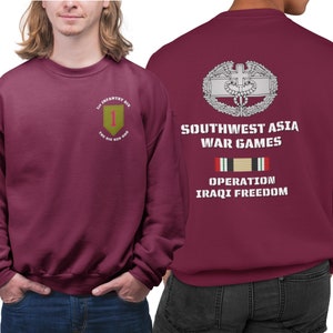 May include: A maroon sweatshirt with a white embroidered patch on the back that says "Southwest Asia War Games" and a ribbon with the text "Operation Iraqi Freedom". The front of the sweatshirt has a patch with the text "1st Infantry Div. The Big Red One" and a number "1" in a yellow square.