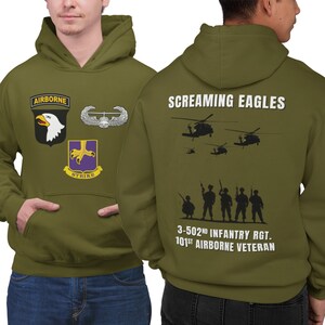 May include: A green hooded sweatshirt with the words "Screaming Eagles" and "3-502nd Infantry Rgt. 101st Airborne Veteran" printed on the back. The front of the sweatshirt has a patch with an eagle and the words "Airborne" and "Strike" printed on it.