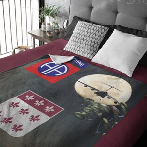 May include: A gray and white fleece blanket with a red, white, and blue patch featuring the 101st Airborne Division insignia. The blanket also features a black and white image of a C-47 airplane dropping paratroopers. The text "ORNE" is visible on the blanket.