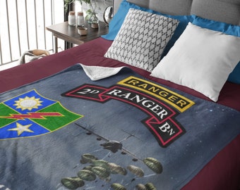 May include: A blue and gray fleece blanket with a military theme. It features a green and yellow patch with a star and lightning bolt, a yellow patch with the text "2nd Ranger Bn", and a silhouette of a plane with parachutes falling from it. The text "And the Way" is visible at the bottom of the blanket.