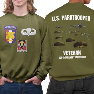 May include: A green sweatshirt with a white print on the back that says "U.S. PARATROOPER VETERAN 509th INFANTRY (AIRBORNE)". The front of the sweatshirt has a patch that says "AIRBORNE" and other patches with military insignia.