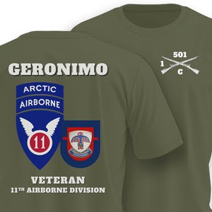 May include: An olive green t-shirt featuring a graphic on the back with the text GERONIMO, ARCTIC AIRBORNE, VETERAN, and 11TH AIRBORNE DIVISION, alongside two military emblems. The front chest area displays the text 501, 1, C with crossed rifles.