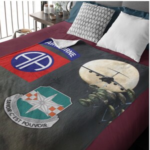 May include: A gray fleece blanket with a red, white, and blue patch with the text "AIRBORNE" and a blue patch with a white "A" inside a circle. The blanket also features a green patch with a white star and the text "SAVOIR C'EST POUVOIR". The blanket has a picture of a plane dropping paratroopers.