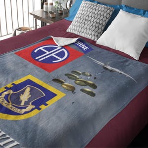 May include: A gray blanket featuring military insignia and a sky scene with parachutes and a plane. The blanket has a red and blue patch with a white emblem, and a yellow and blue patch with text. The word "AIRBORNE" is visible.