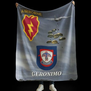 May include: A blanket featuring military-themed graphics. The design includes a red and yellow lightning bolt emblem, a blue and red crest with a white eagle, and the word "GERONIMO" in white. A plane and parachutes are also depicted.