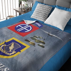 May include: A gray blanket featuring military insignia and a sky scene with parachutes and an airplane. The blanket has a red and blue patch with the text "AIRBORNE" and a yellow and blue patch with the text "STRIKE HOLD".