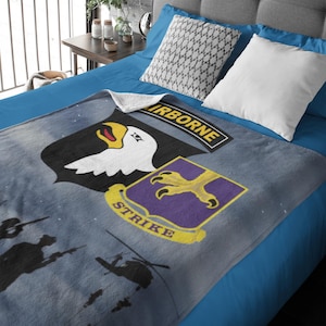 May include: A gray blanket featuring military insignia, including a black shield with a white eagle head and the word "AIRBORNE". A purple shield with a yellow claw and the word "STRIKE" is also visible. Silhouettes of soldiers and a helicopter are in the background.