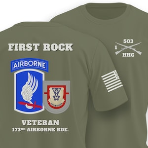 May include: An olive green t-shirt featuring the text FIRST ROCK and VETERAN 173RD AIRBORNE BDE. on the back, with the 173rd Airborne Brigade insignia. The front chest displays 503 1 HHC with crossed rifles, and a white American flag on the sleeve.