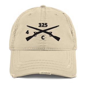 May include: A beige distressed baseball cap with a black embroidered design featuring crossed rifles, the number "325", and the letter "C".