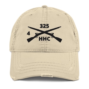 May include: A distressed beige baseball cap with a curved brim. The cap features a black graphic of crossed rifles with the numbers "325" and "4" above and below the rifles, and the letters "HHC" below.