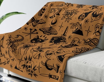 May include: A brown throw blanket with a black and white pattern of various tattoo-style designs, including skulls, hearts, stars, and a crescent moon. The blanket is draped over a white couch.