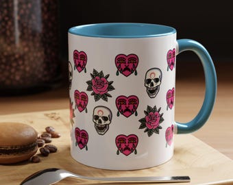 May include: White ceramic mug with a blue interior and handle, decorated with pink hearts, roses, and skull illustrations. A spoon and a macaron are in the foreground. The mug is on a wooden surface.